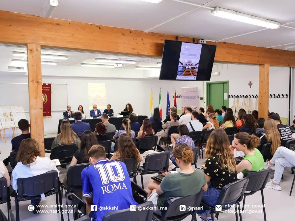 Pamje e gjerë e një salle konference me pjesëmarrës dhe një panel folësish përpara flamujve.
